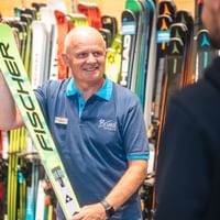 Manfred Rogetzer steht in Bründl Sports Kleidung vor einer Wand mit Skiern, hält ein Paar Fischer-Ski in der Hand und spricht mit einer Person im Vordergrund.