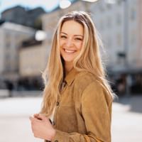 Paula Bründl stands smiling with a shopping bag in a sunny city square. Light-colored buildings are visible blurred in the background.