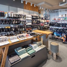 Das Bild zeigt einen Verkaufsbereich in einem Bründl Sports Shop mit einer großen Auswahl an Handschuhen, Mützen und Wintersportaccessoires, die an den Wänden und Ständern präsentiert werden. In der Mitte stehen Tische mit gefalteten T-Shirts und Hoodies.