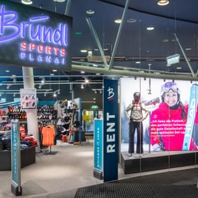 Das Bild zeigt den Eingang eines Bründl Sports Shops in der Planai mit einem großen, leuchtenden Bründl Sports Schild. Im Inneren sind verschiedene Wintersportprodukte wie Kleidung und Ausrüstung ausgestellt, und es gibt einen Bereich für Verleih und Service. Ein großes Werbeposter zeigt eine lächelnde Skifahrerin.