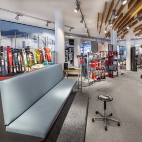 Das Bild zeigt eine Skiausrüstungs-Abteilung mit einer Auswahl an Skiern, Stöcken und Skischuhen, sowie einer Sitzgelegenheit für Kunden, um Schuhe anzuprobieren.