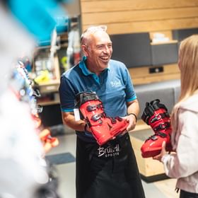 Ein Bründl Sports Mitarbeiter berät eine Kundin bei der Auswahl von Skischuhen. Beide halten rote Skischuhe in der Hand und lachen, während sie sich im Shop umsehen.