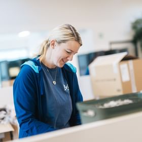 A woman stands at a worktable handling small items in a box or on a tray. She smiles while looking down, with boxes and materials visible in the background.