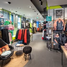 Das Bild zeigt den Innenbereich eines Bründl Sports Shops, in dem farbenfrohe Winter- und Outdoor-Bekleidung sowie Accessoires verschiedener Marken ordentlich auf Ständern und Tischen ausgestellt sind.
