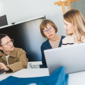 Three women are standing and sitting at a table with open laptops, talking to each other. One woman looks at the others while an older woman in the middle speaks and the third listens attentively.