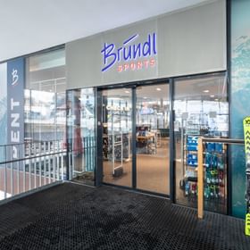 Das Bild zeigt den Eingang eines Bründl Sports Shops mit Glastüren und beleuchtetem Logo. Im Inneren sind Skiausrüstung und Zubehör sichtbar, während vor dem Eingang zwei Skier aufgestellt sind.