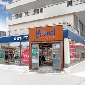 The image shows the entrance of a sports store with the sign “Bründl Sports” above the door. Large display windows show mannequins, and the words “OUTLET” and “SALE” are clearly visible.