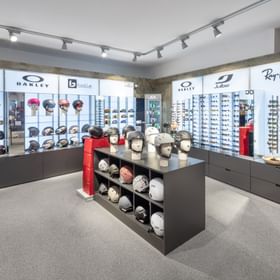 Das Bild zeigt eine Präsentation von Skihelmen und Sonnenbrillen bekannter Marken wie Oakley, POC, und Ray-Ban in einem gut beleuchteten Ausstellungsbereich eines Sportgeschäfts.