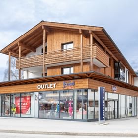 Das Bild zeigt das Bründl Sports Outlet-Geschäft in einem modernen Gebäude im alpinen Stil mit viel Holz, großen Fenstern und einem klaren "Outlet"-Schild an der Fassade.
