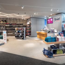 Das Bild zeigt den Innenbereich eines Bründl Sports Shops mit einer großen Auswahl an Skischuhen, Helmen und Zubehör. Auf einem Tisch sind gefaltete Kleidungsstücke gestapelt. Rechts ist ein Verkaufs- und Servicebereich mit einer Holztheke und einer Bildschirmanzeige zu sehen.
