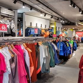 Interior view of the Bründl Sports Outlet in Kaprun featuring a wide selection of sportswear, jackets, shirts and pants in various colors. Discounted clothing is displayed on racks and wall shelves, with red sale signs advertising discounts of up to 50%.