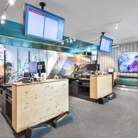 Das Bild zeigt den Verleihbereich eines Bründl Sports Shops, mit Holzcountern, Bildschirmen zur Buchungsinformation und großen Fotomotiven von Outdoor-Sportarten. Im Hintergrund befindet sich eine Sitzbank mit einem großen Bildschirm, der Wintersportbilder zeigt.