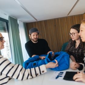 Four people stand around a table examining a blue jacket together. They hold the garment and look at it while discussing it.