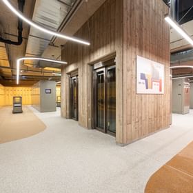 The image shows a modern ski depot area inside the Bründl Sports AreitXpress valley station shop with yellow, white, and orange lockers and elevators in the center. A wayfinding map with depot numbers is mounted on the wall, including “001–174,” “175–358,” and “477–496.”