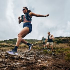 Trailrunner springen über eine matschige Passage auf einem Bergtrail und tragen Hoka Trailrunning-Schuhe.