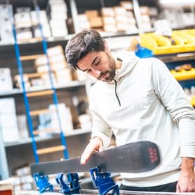 Ein Mann überprüft die Lauffläche eines Skis in einer Werkstatt, umgeben von Werkzeugen und Regalen mit Material.