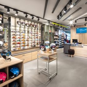 Das Bild zeigt den Innenbereich eines Bründl Sports Shops mit einer großen Auswahl an Helmen und Skibrillen, die in Regalen präsentiert werden, sowie einem Empfangsbereich mit einer Kasse im Hintergrund.