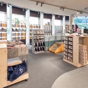The image shows a bright retail area inside the Bründl Sports AreitXpress valley station shop with shelves full of hiking shoes, sports shoes, and shoe boxes. Wooden display units with more shoes and accessories are placed throughout the room, trekking poles and bags are visible on the left, and the “On” logo can be seen on several boxes.