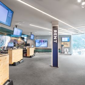 Das Bild zeigt den Verleihbereich eines Bründl Sports Shops mit Check-in-Stationen, Holztheken und mehreren Bildschirmen. Eine Säule mit der Aufschrift "RENT" ist zentral platziert. Im Hintergrund ist eine Sitzgelegenheit zu sehen.