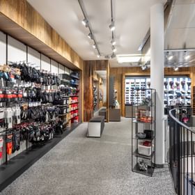 Zu sehen ist ein Verkaufsbereich im Shop Bründl Sports Saalbach life.style mit vielen Handschuhen, Socken und Wintersport-Zubehör an der linken Wand. Im Hintergrund sind Regale mit Skihelmen und Skibrillen zu sehen, rechts befindet sich ein Treppenabgang mit Geländer.