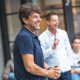 The image shows Bernhard Bründl wearing a dark blue polo shirt, laughing in a relaxed conversation. In the background, Christoph Bründl is visible in a white shirt, also gesturing and smiling, while other people attentively observe the discussion.