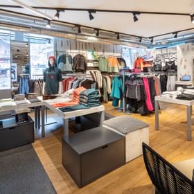 Das Bild zeigt einen Verkaufsbereich in einem Bründl Sports Shop mit verschiedenen Winter- und Outdoor-Kleidungsstücken, die auf Tischen und Kleiderständern präsentiert werden. Schaufensterpuppen tragen Freizeitmode, und im Vordergrund steht eine Sitzgelegenheit, was eine einladende Einkaufsatmosphäre schafft.
