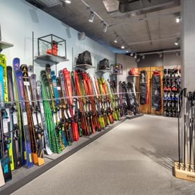 Das Bild zeigt eine Verkaufsfläche in einem Bründl Sports Shop mit einer breiten Auswahl an Skiern in verschiedenen Farben und Marken. Daneben sind Skistöcke ausgestellt, und im Hintergrund gibt es Regale mit Skischuhen und Taschen.
