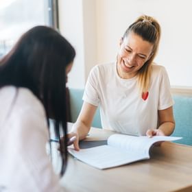 Two women are sitting at a table in a bright room. One woman is smiling and pointing at an open document, while the other is looking down and writing.
