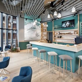 Modern café with wooden interior, large windows, lounge chairs, and turquoise bar area.