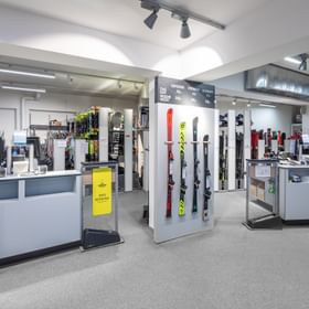 Das Bild zeigt den Ski-Verleihbereich eines Ladens, mit einer Ausstellung von verschiedenen Skimodellen und einer Preistafel. Im Hintergrund sind zahlreiche Skier und Zubehör sichtbar, sowie ein Counter für Beratung und Buchung.