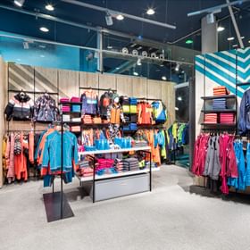 Das Bild zeigt einen farbenfrohen Verkaufsbereich eines Bründl Sports Shops mit Winterbekleidung, Jacken und Pullovern, die auf Regalen und Kleiderständern ausgestellt sind. Rucksäcke und Accessoires hängen an der Seite, und die Kleidung ist ordentlich nach Farben und Stilen sortiert.