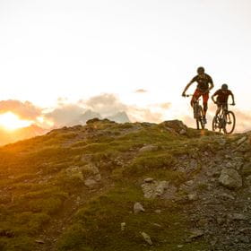 two mountainbikers are riding through the mountains, while the sun sets in the background