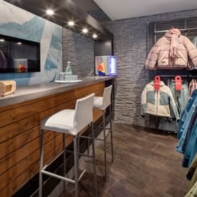The image shows the interior of a shop with a wooden counter and two tall chairs on the left side. On the right, several winter jackets are displayed with red “SALE” tags, and on the counter there are glasses, a bottle, and a small refrigerator.
