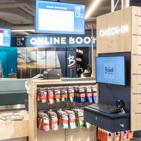 The image shows a check-in terminal inside the Bründl Sports AreitXpress valley station shop with a screen, keyboard, scanner, and a height scale from 1 m to 2 m on the right side. Accessories such as socks are visible in the background, along with the texts “CHECK-IN,” “SHOP BOOKING,” “RENT BOOTS,” and “ONLINE BOOT.”