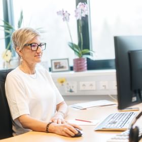 A woman wearing glasses sits at a desk using a computer mouse in front of a monitor. A keyboard, a pen, and papers are on the desk, and plants are visible on the windowsill in the background.
