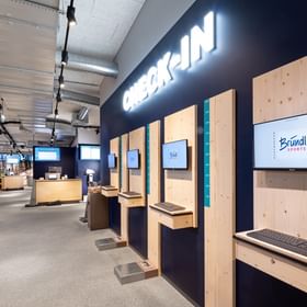 Zu sehen ist der Check-in Bereich im Shop Bründl Sports Penkenbahn Talstation Mayrhofen mit mehreren Holz-Terminals, Bildschirmen, Tastaturen und Größenmessungen. Auf der dunklen Wand steht groß der Text „CHECK-IN“, auf mehreren Bildschirmen ist das Bründl Sports Logo sichtbar.