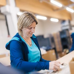 A woman stands at a worktable placing an item into a bag or packaging. In the background, there are boxes and another person working.