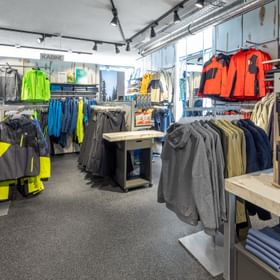 Das Bild zeigt eine Verkaufsfläche in einem Bründl Sports Shop, auf der eine große Auswahl an Winterjacken, Hosen und Outdoor-Bekleidung in verschiedenen Farben präsentiert wird. An der Wand befindet sich ein Schild für Umkleidekabinen, und Accessoires wie Gürtel sind ebenfalls zu sehen.