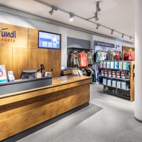 Das Bild zeigt den Empfangs- und Kassenbereich eines Bründl Sports Ladens, mit einem Holz-Counter, einem Bildschirm an der Wand und Kleidungsstücken sowie Zubehör in Regalen im Hintergrund.