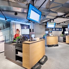 Zu sehen ist ein Skiverleih- und Buchungsbereich im Shop Bründl Sports Penkenbahn Talstation Mayrhofen mit mehreren Holztheken, Computern, Kartenlesern und Bildschirmen. 