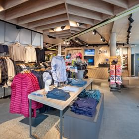 Das Bild zeigt einen Bründl Sports Shop mit einer Vielzahl an Winterkleidung, Mützen und Schuhen. Die helle, moderne Einrichtung mit Holzdetails vermittelt eine warme und einladende Atmosphäre.
