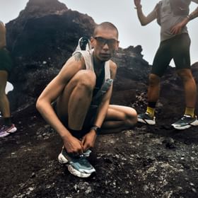 Trailrunner bindet seine Hoka Trailrunning-Schuhe auf felsigem Untergrund, während zwei weitere Läufer im Hintergrund stehen.