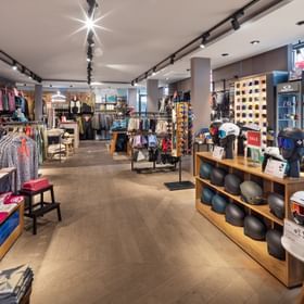 The image shows a large retail space with clothing displayed on tables and racks, as well as many helmets on shelves in the foreground. Several red signs with the word “SALE” are visible, and on the right wall additional helmets and goggles are displayed.