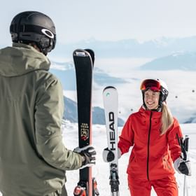 Skier in a green outfit talking to a woman in red ski clothing holding Head skis on a snowy slope with mountain panorama.