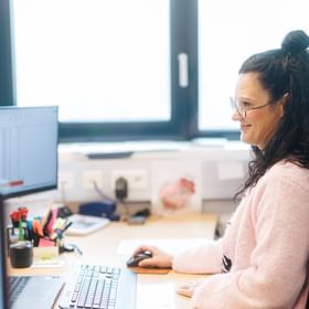 A woman wearing glasses sits at a desk looking at a computer screen displaying tabular data. A keyboard, mouse, and office supplies are on the desk, with windows visible in the background.
