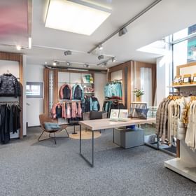 Zu sehen ist ein heller Verkaufsbereich im Shop Bründl Sports Saalbach life.style mit Winterjacken, Hosen, Westen und Lifestyle-Bekleidung auf Kleiderstangen und Wandregalen. Rechts ist der Schriftzug „Luhta“ sichtbar, in der Mitte stehen ein Tisch, Sessel und kleine Hinweisschilder.