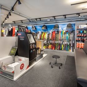 Das Bild zeigt einen Bereich in einem Bründl Sports Shop mit einer Boot-Fit-Station zur individuellen Anpassung von Skischuhen, umgeben von Regalen mit Skiern, Skischuhen und Zubehör. Es gibt auch eine Sitzbank und einen Hocker, die für eine komfortable Anprobe vorgesehen sind.