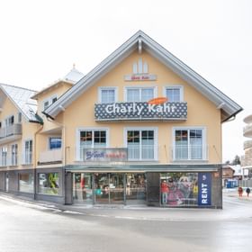 Das Bild zeigt ein Gebäude mit dem "Bründl Sports" Shop im Erdgeschoss und einem Schild für "Charly Kahr" im oberen Bereich, umgeben von anderen Gebäuden in einer winterlichen Umgebung.