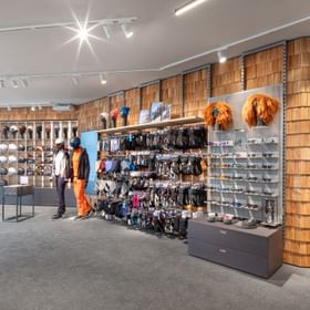Das Bild zeigt eine Bründl Sports Shop-Ecke mit einer großen Auswahl an Skihelmen, Handschuhen, Brillen und anderem Zubehör. Zwei Schaufensterpuppen in Winterkleidung stehen als Dekoration. Rechts ist eine Trinkstation zu sehen, die Kunden zur Vermeidung von Plastik nutzen können