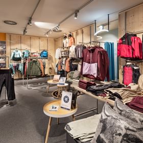 The image shows a retail area inside the Bründl Sports AreitXpress valley station shop with jackets, pants, shirts, and outdoor clothing displayed on racks, wall shelves, and tables. Folded clothing and a large patterned cushion are visible in the foreground, with small information signs placed on a table.